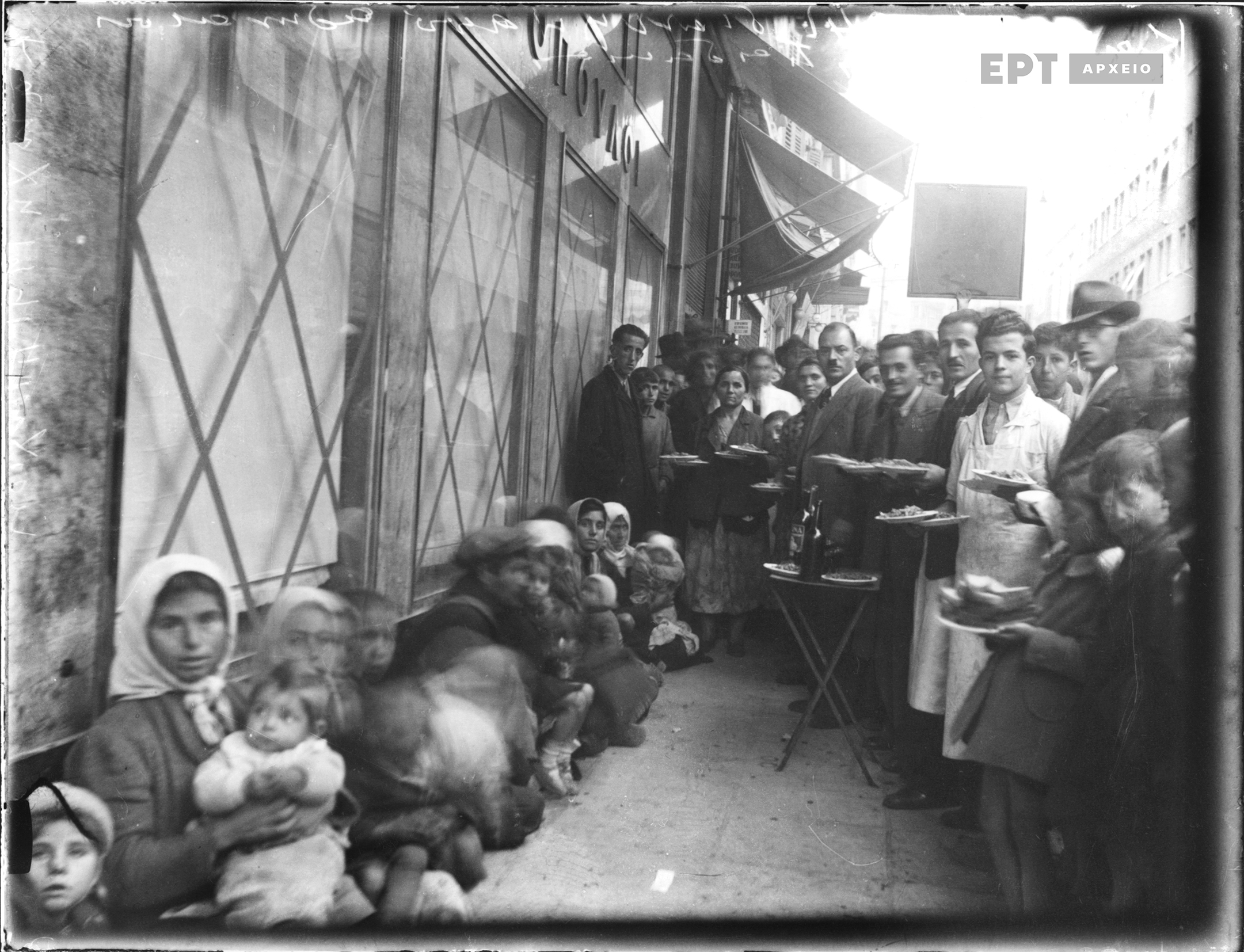 Αθήνα. Χαυτεία. Πρόσφυγες καθισμένοι έξω από το κατάστημα Αφοί Λαμπρόπουλοι περιμένοντας να πραγματοποιηθεί η διανομή συσσιτίου. Πολίτες κρατούν στα χέρια τους πιάτα με φαγητό και ψωμί. Πλήθος κόσμου είναι συγκεντρωμένο. Συλλογή Πέτρου Πουλίδη, Φωτογραφικό Αρχείο ΕΡΤ.